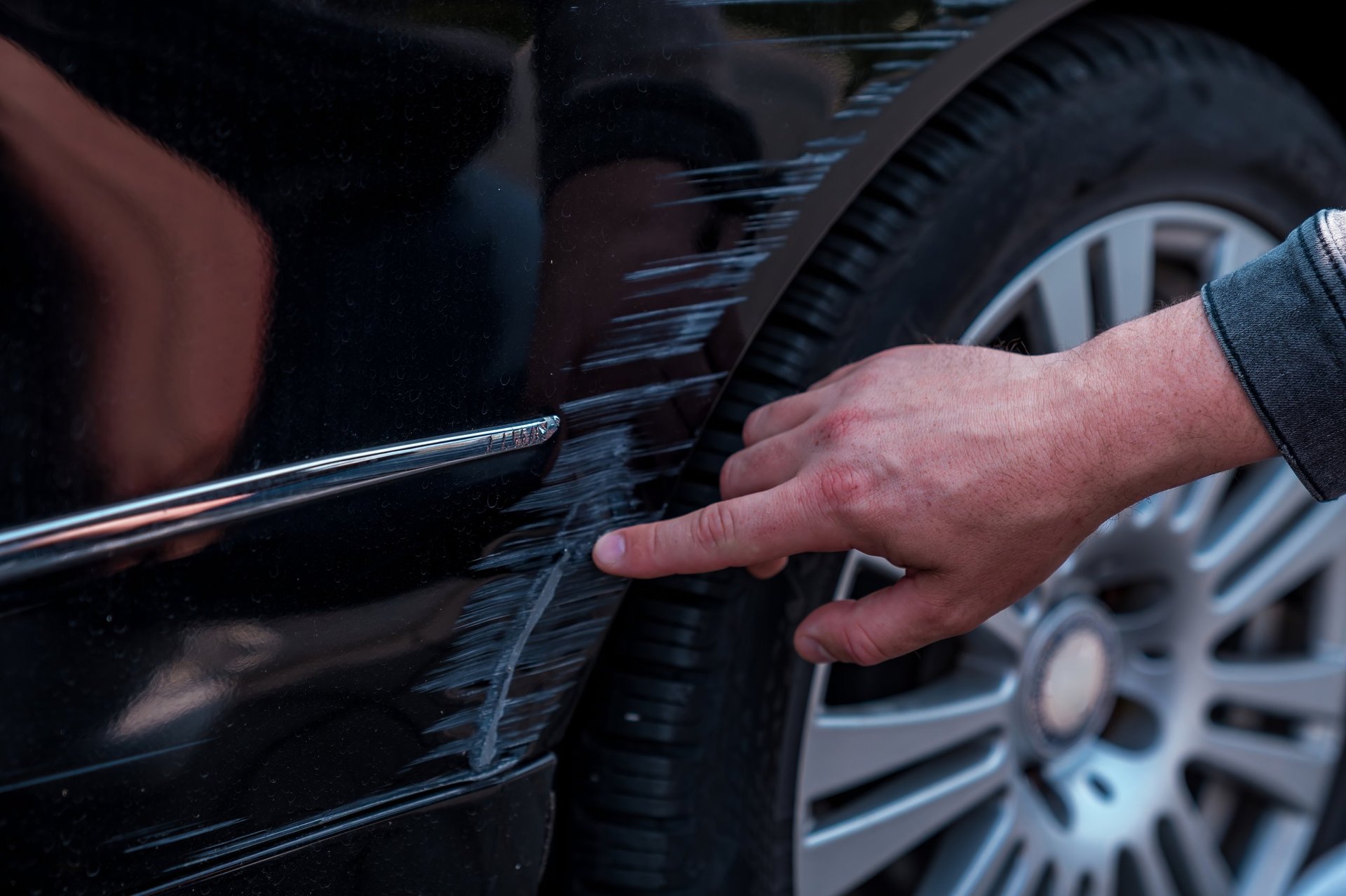 Man inspecting a fresh scratch on the side of a black car, examining vehicle damage for insurance claims
