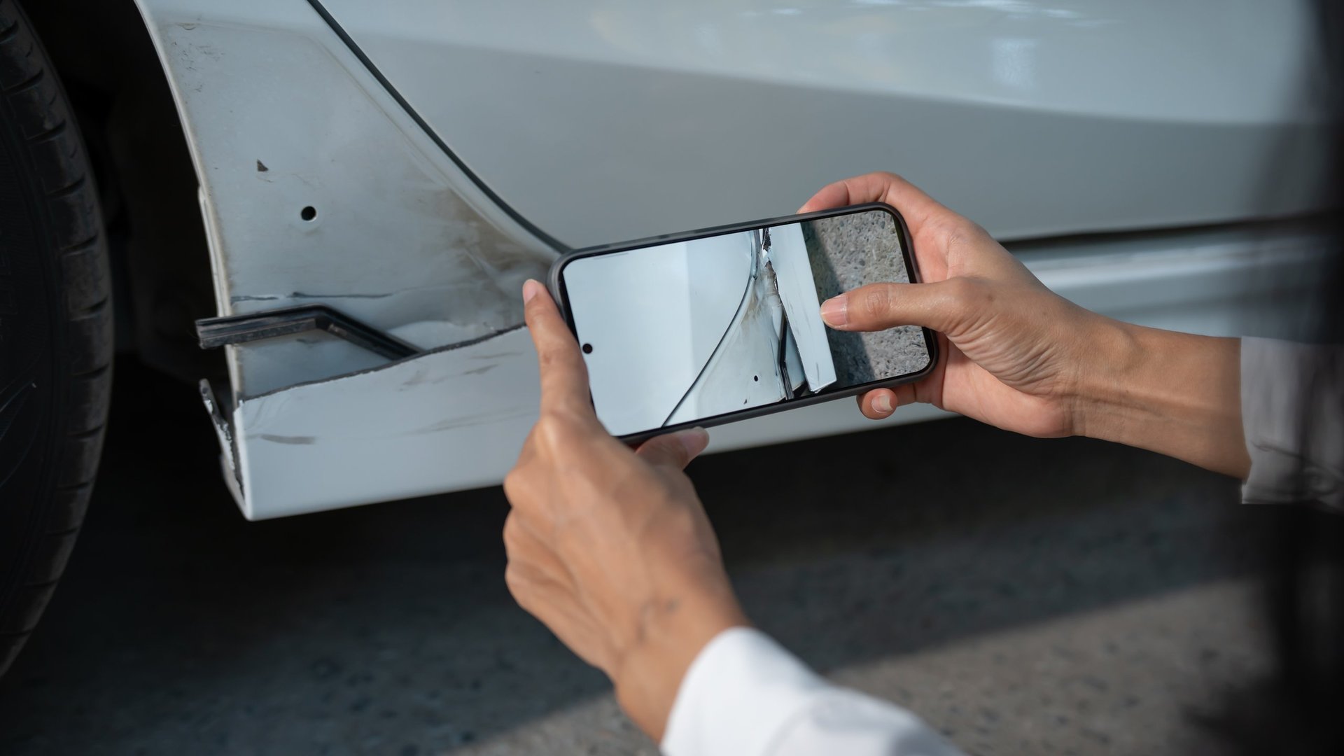 Person using smartphone to photograph car body damage and dent on a white vehicle, documenting accident for insurance claim or repair estimate.