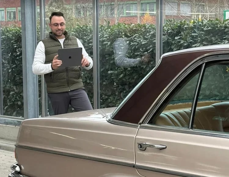 Man in olive vest holding tablet standing next to vintage beige car in modern garage with glass windows