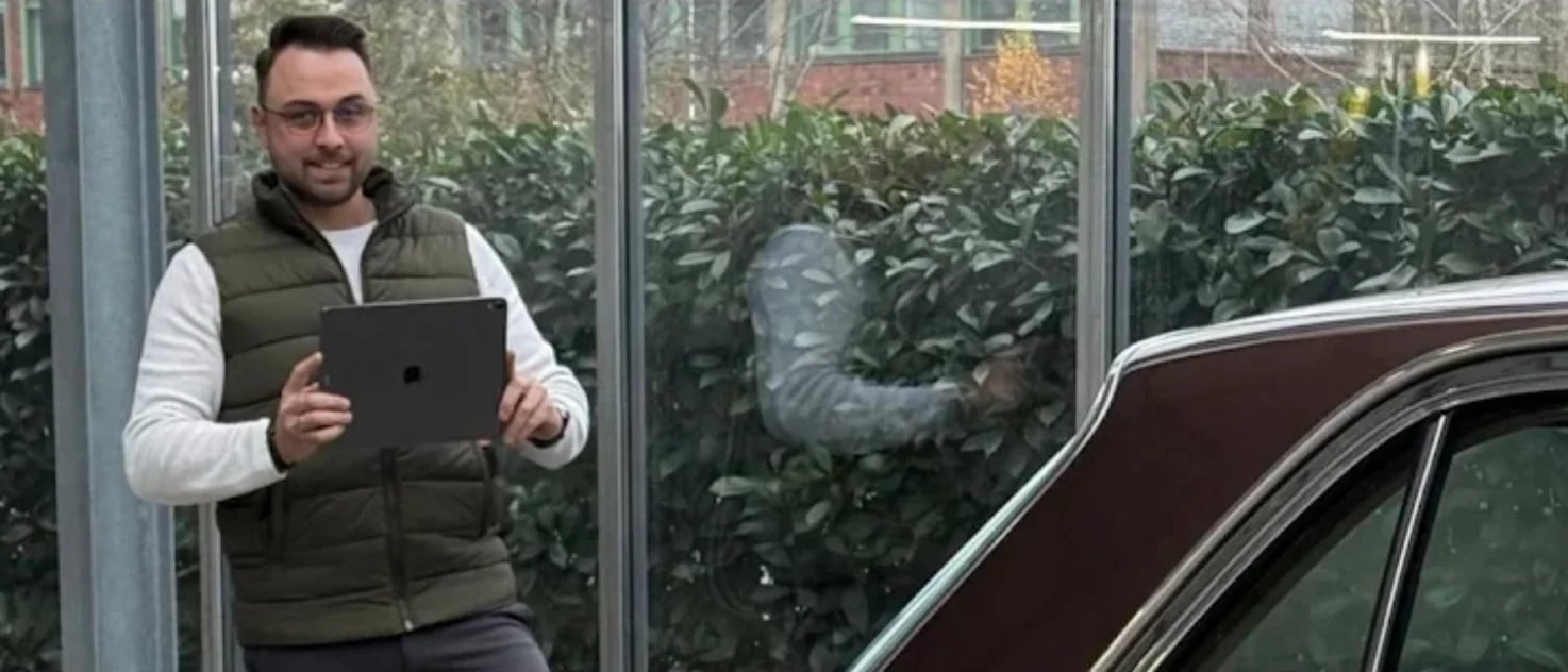 Man holding tablet standing indoors next to vintage car with greenhouse windows behind him