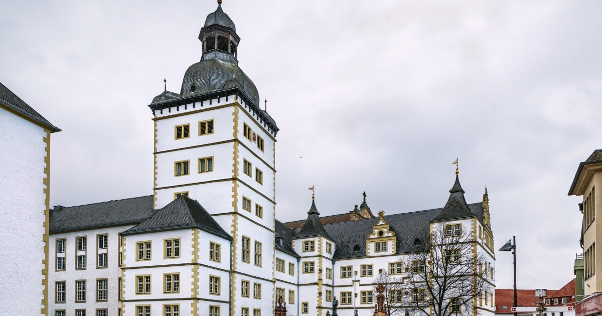 Historic European castle or mansion with white and cream colored stone walls, multiple turrets with dark spires, and traditional architecture against a cloudy sky