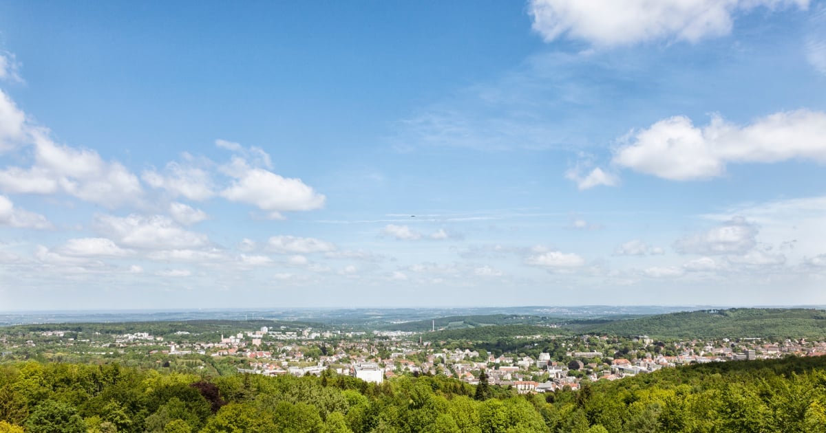 Aerial view of a suburban town nestled in a valley surrounded by green forested hills under a partly cloudy blue sky