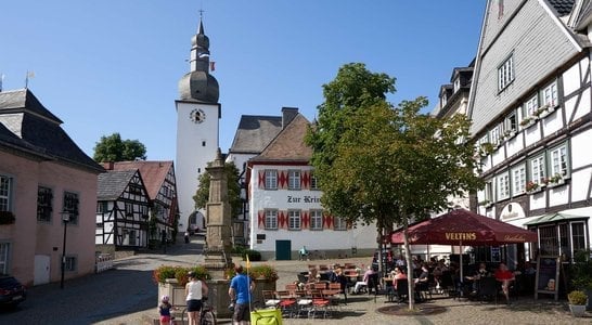 Historic European town square with half-timbered buildings, clock tower, outdoor café seating, and blue sky